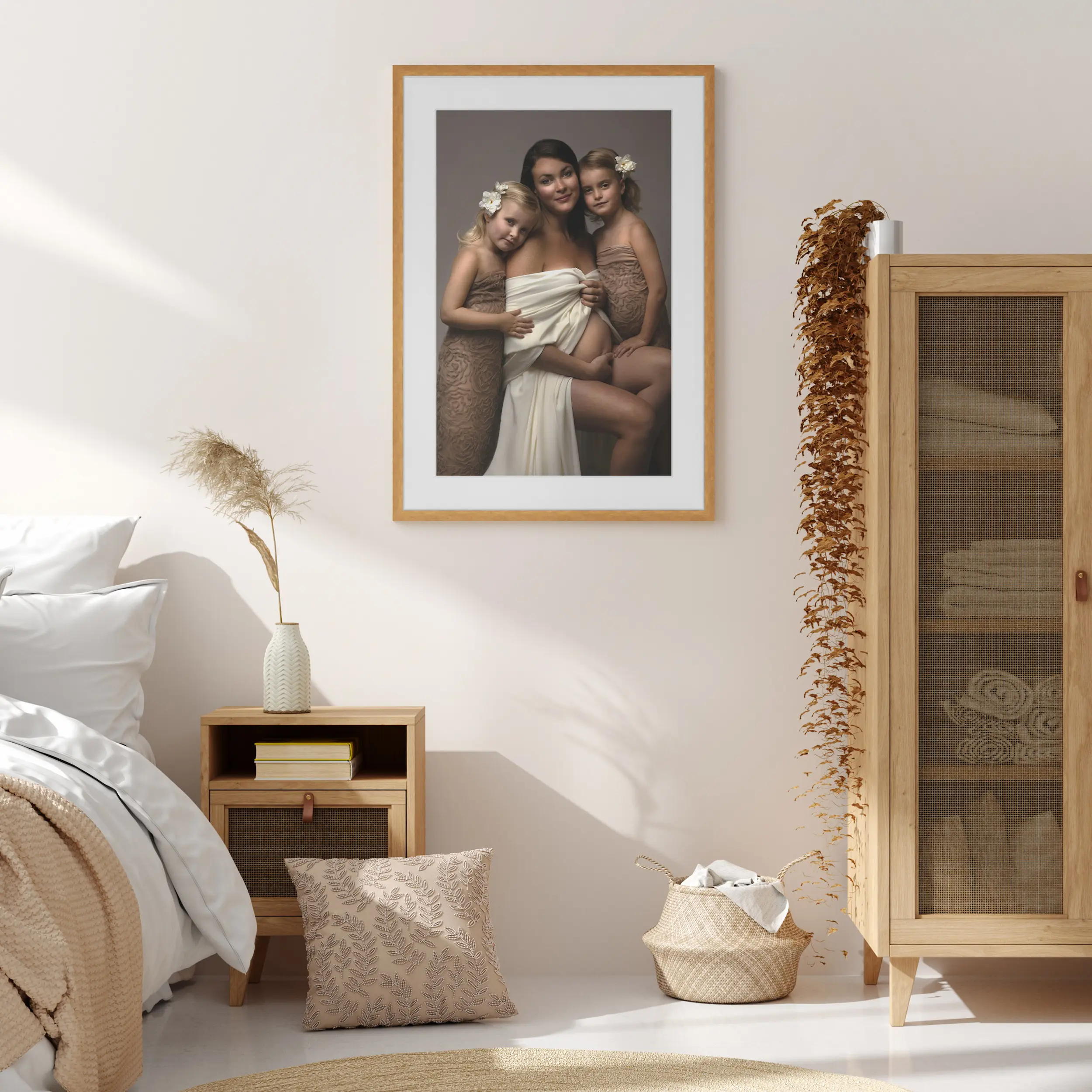 Framed maternity portrait of a mother with two daughters displayed in a soft, natural-toned bedroom with warm wood furniture