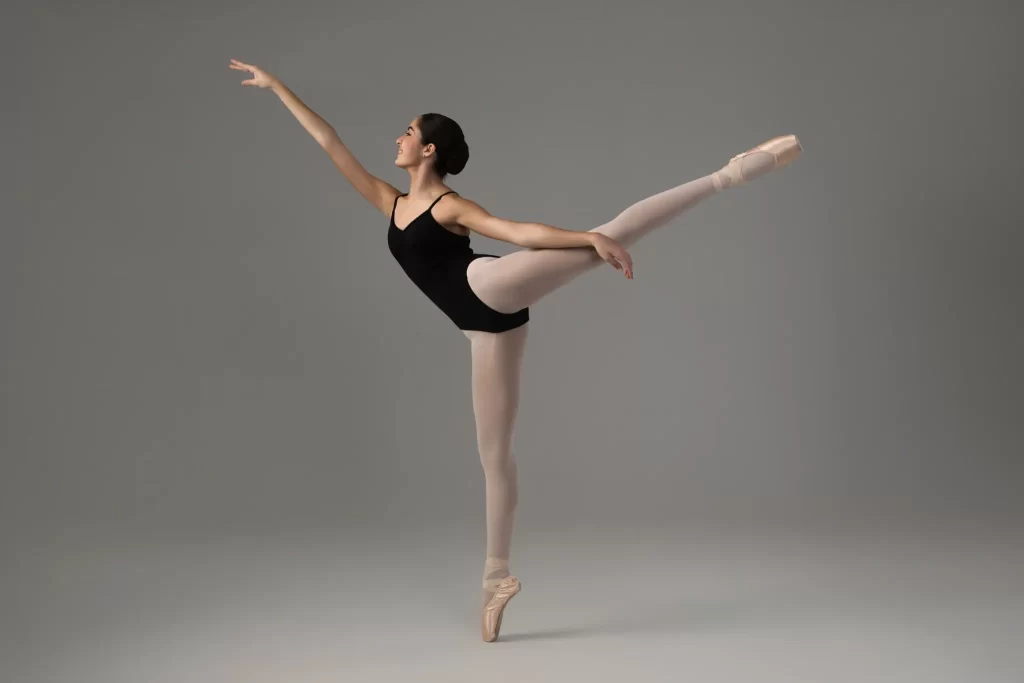 Ballet dancer in black leotard and pink tights holding an arabesque on pointe in a studio setting