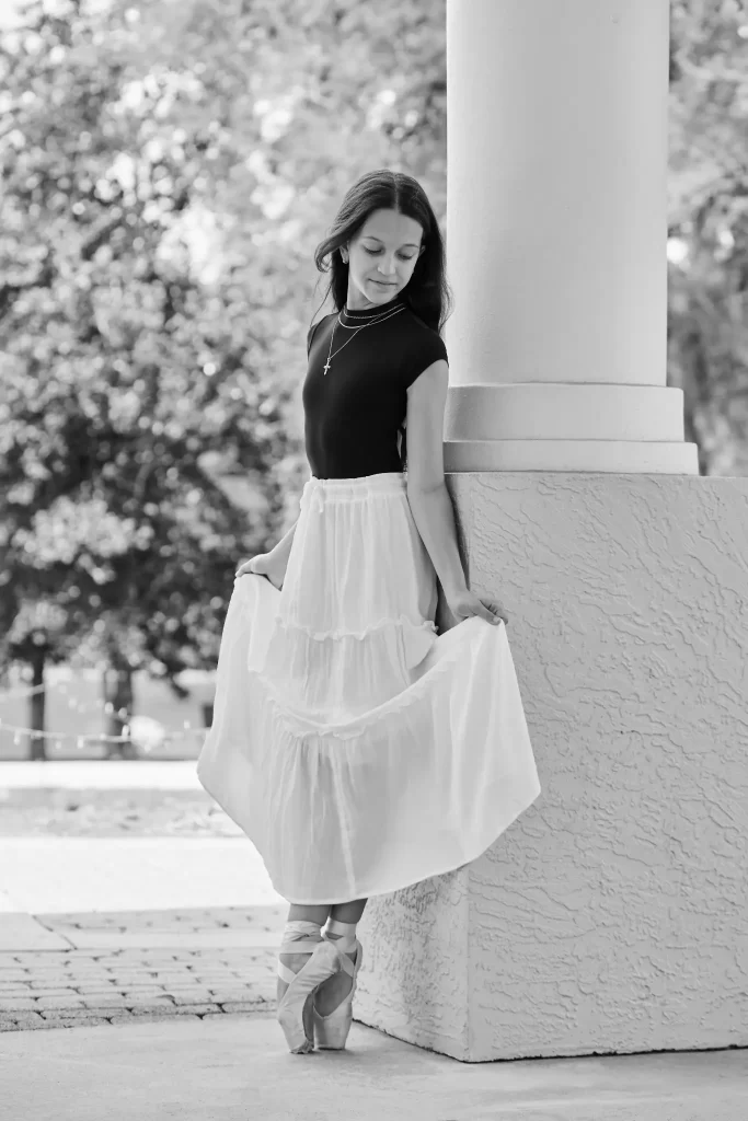 Ballet dancer in a white skirt and black top standing en pointe beside a column in a sunlit outdoor setting