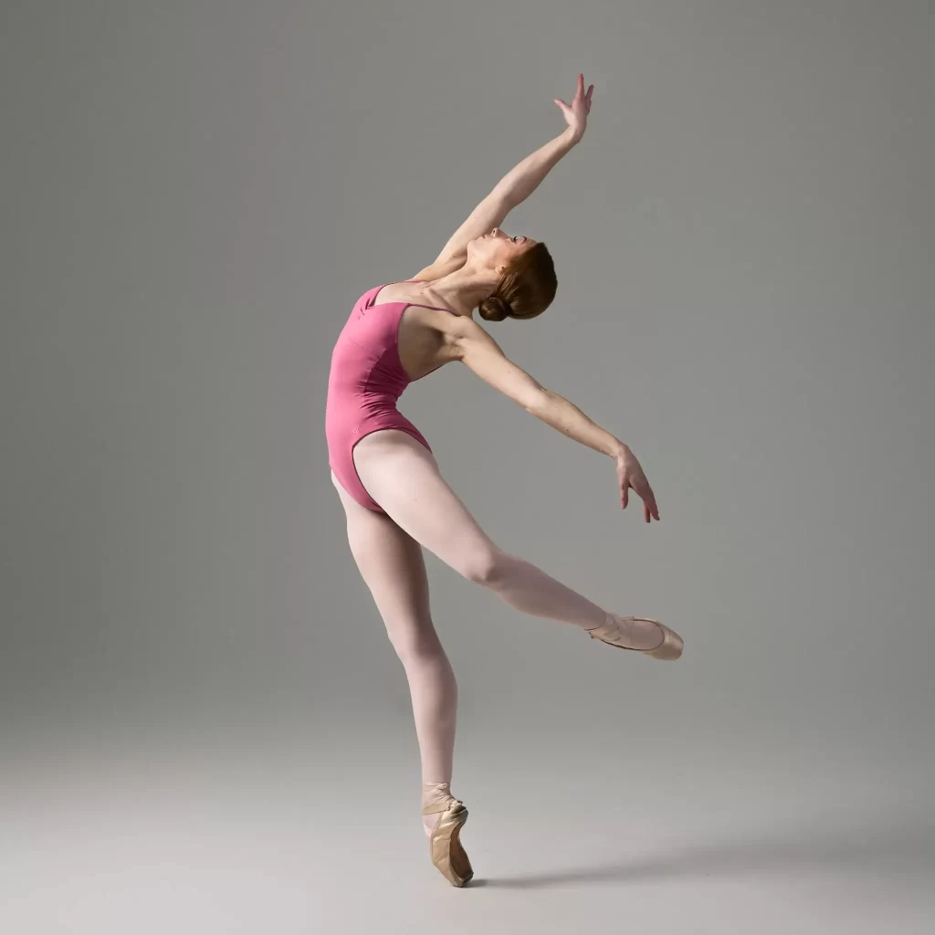 Ballet dancer in a pink leotard performing an extended pose on pointe against a gray background