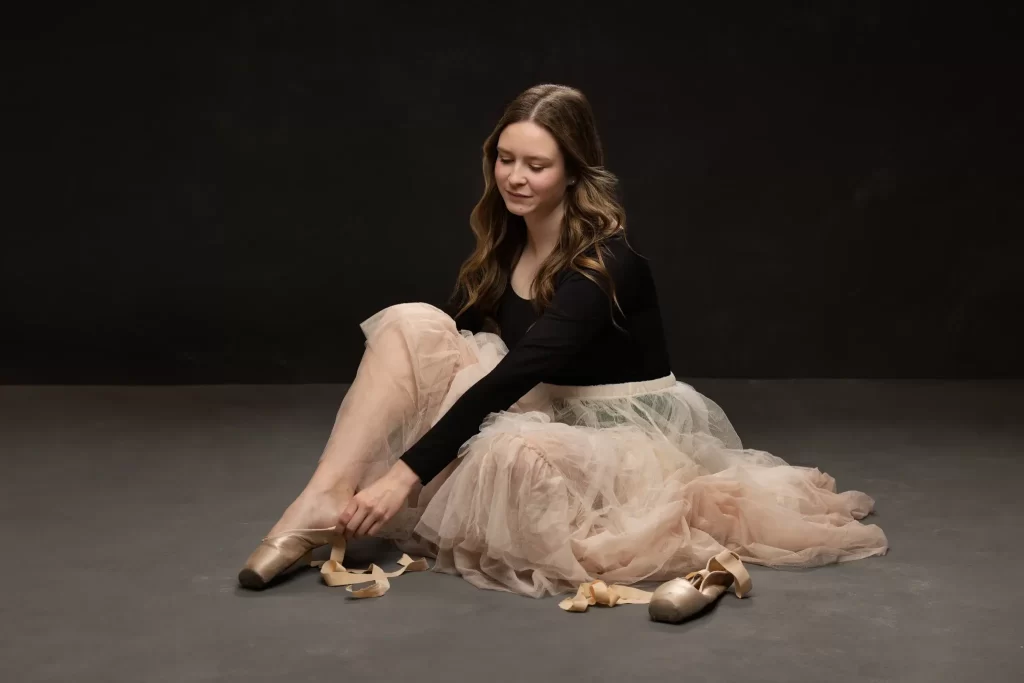 Dancer in a soft tulle skirt tying her pointe shoes while seated on the studio floor