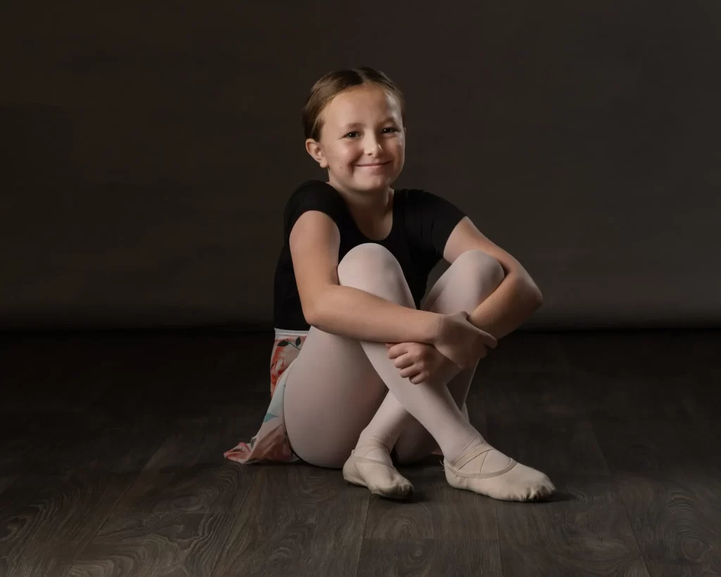 Young ballet student in black leotard sitting on the floor with a soft smile against a dark backdrop