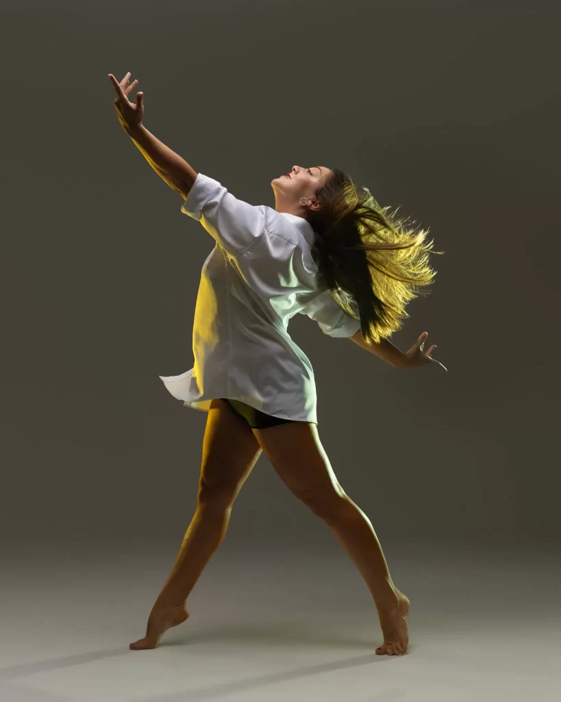 Dancer backlit in a white button-down shirt with hair in motion and arms extended