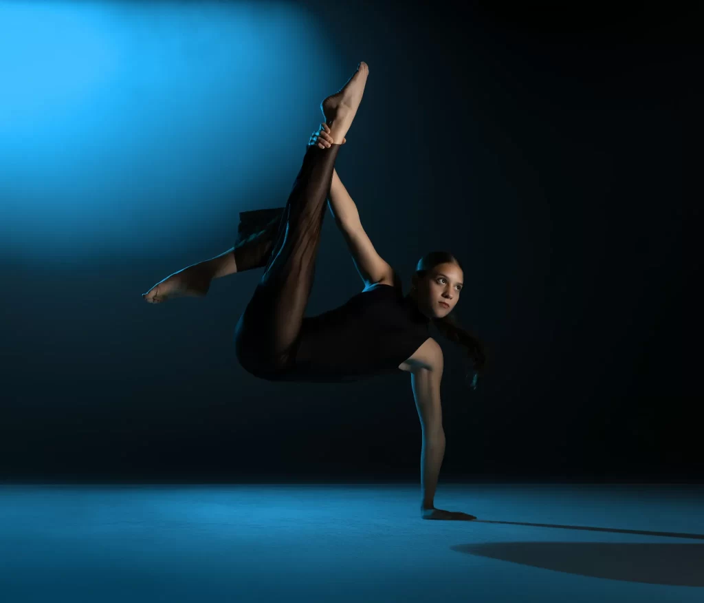 Female dancer in black costume holding a leg extension on one hand under dramatic blue studio light