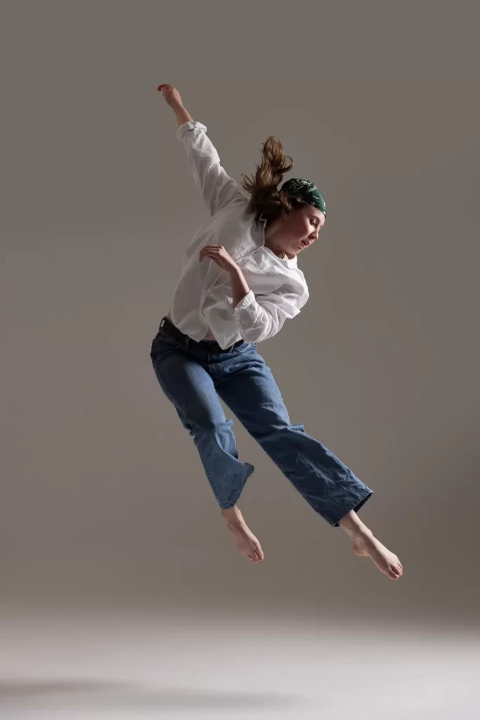 Dancer in jeans and white shirt mid-air against a neutral gray studio backdrop