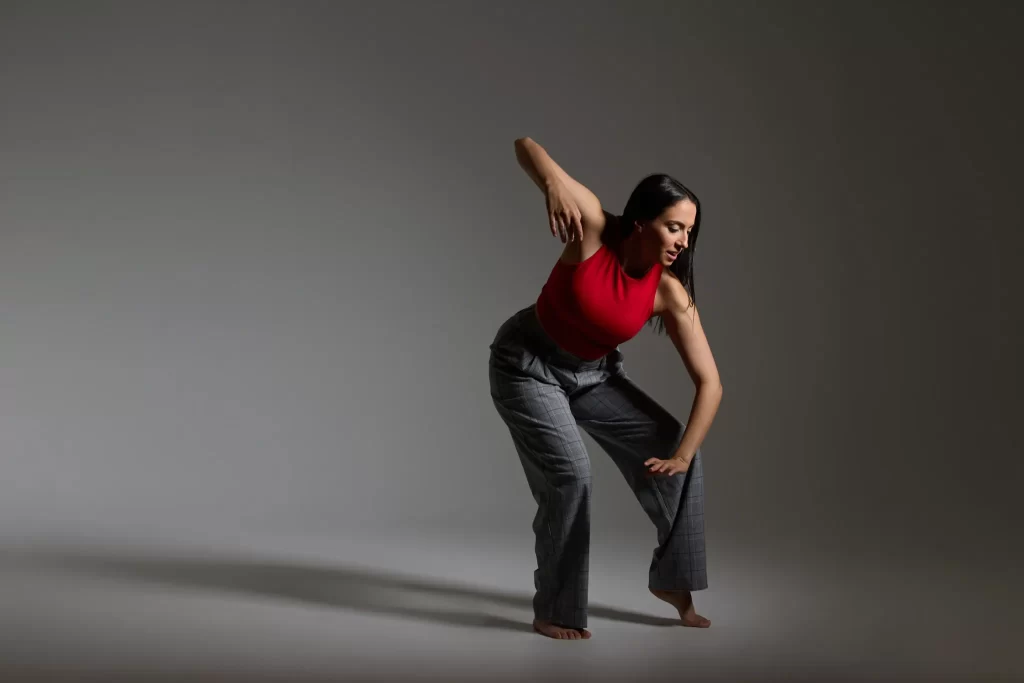 Female dancer in red top and gray plaid pants performing a contemporary pose against a neutral background