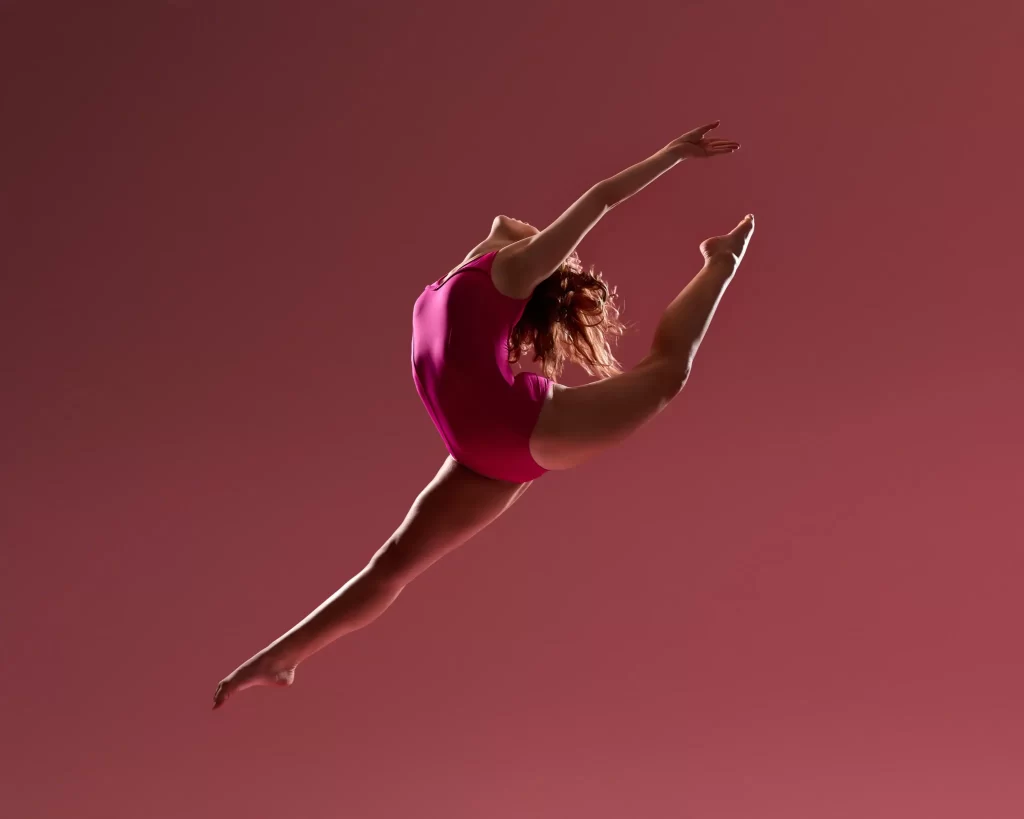 Dancer mid-leap in a magenta leotard performing a split jump against a soft red background