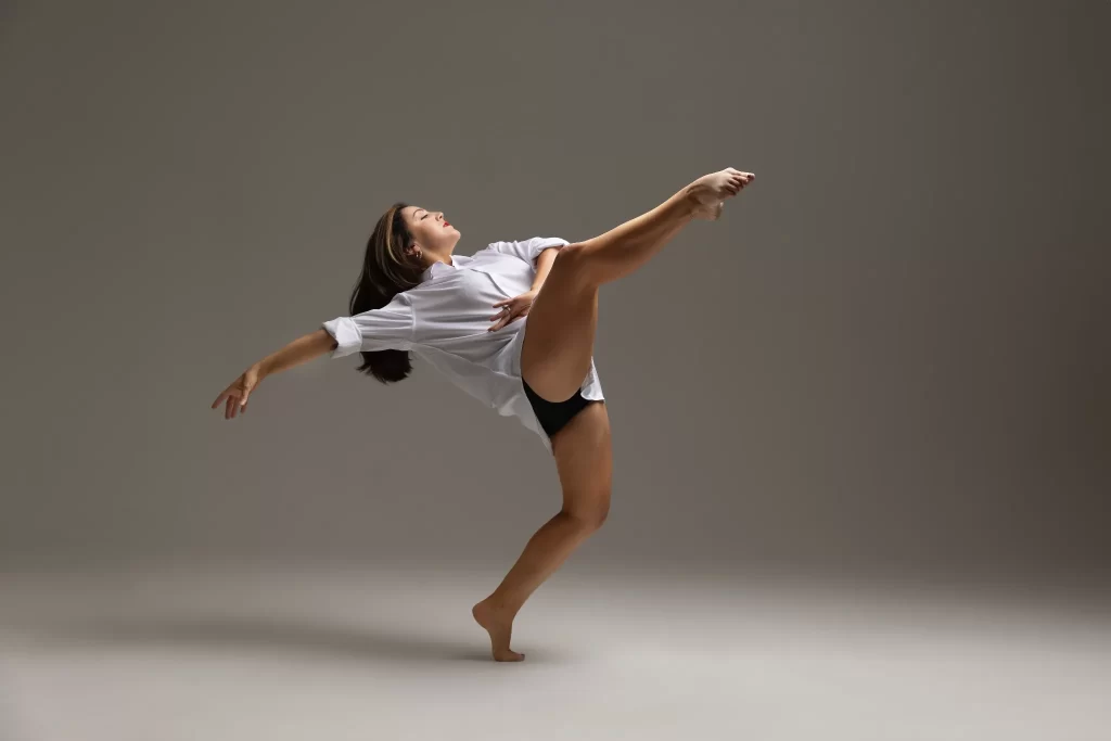 Contemporary dancer in white shirt and black shorts performing a high kick against a gray backdrop